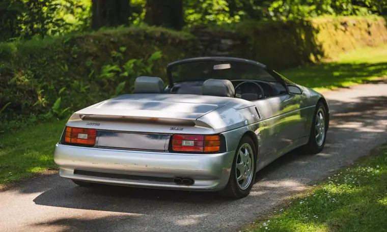 Vista tre quarti posteriore del prototipo Lotus Elan M100 del 1986 di colore argento parcheggiato su una strada sterrata immersa verde.